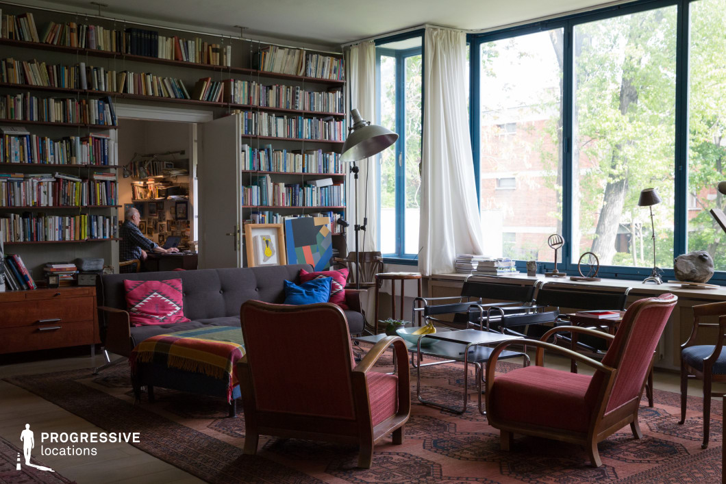 This sophisticated living space blends mid-century modern aesthetics with a literary ambiance, featuring a floor-to-ceiling bookshelf packed with an extensive collection of books. The room is illuminated by large blue-framed windows, allowing ample natural light to enhance the warm tones of the vintage wooden furniture. A sleek black sofa with vibrant throw pillows and a patterned blanket sits alongside deep red upholstered armchairs, creating a comfortable yet structured seating arrangement. The decor incorporates Bauhaus-style elements, such as metal-framed coffee tables and minimalist sculptural objects displayed on the window ledge. A vintage industrial floor lamp adds an artistic touch, while layered Persian rugs introduce texture and warmth. This environment is ideal for intellectual, creative, or period-piece settings, offering a refined yet inviting backdrop for storytelling and film production.