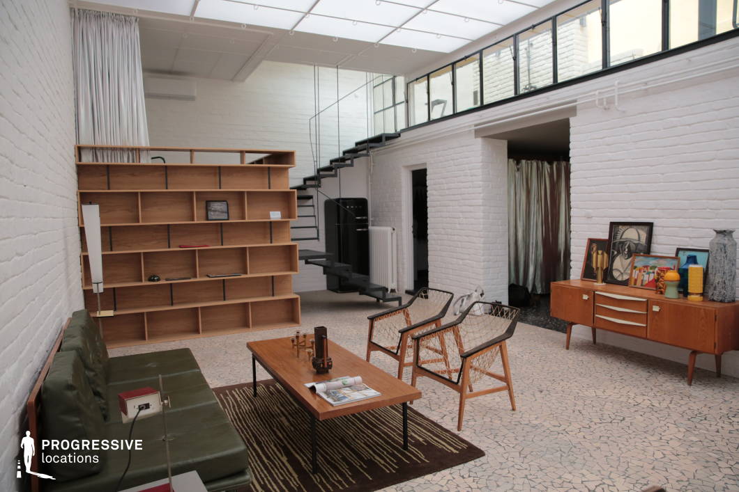 This open-concept loft blends mid-century modern design with industrial influences, featuring white-painted exposed brick walls and a striking glass ceiling that floods the space with natural light. A sleek black metal staircase leads to an upper mezzanine level, adding a dynamic architectural element. The room is furnished with a mix of vintage and contemporary pieces, including a low-profile olive-green sofa, woven lounge chairs, and a wooden coffee table atop a geometric-patterned rug. A modular wooden bookshelf with an asymmetrical design serves as both a divider and a display space. A mid-century sideboard against the wall holds abstract artwork, sculptural vases, and decorative objects, reinforcing the artistic and curated aesthetic. The combination of textures, from the terrazzo-style flooring to the metallic curtain, creates a layered, sophisticated atmosphere, making this an ideal location for editorial shoots, design showcases, or lifestyle productions.