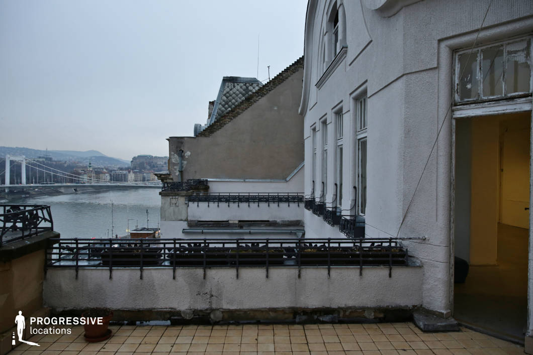 This elevated terrace offers a stunning panoramic view of a historic cityscape, overlooking a river with an elegant suspension bridge and rolling hills in the background. The building’s façade exhibits a mix of classic and Art Nouveau influences, featuring curved window frames, decorative wrought-iron railings, and sculptural elements. The aged exterior, with peeling paint and worn surfaces, adds a sense of history and character. The tiled terrace floor, accompanied by an empty planter, enhances the abandoned yet cinematic atmosphere. An open doorway leads into a dimly lit interior space, hinting at untold stories within. This location is ideal for atmospheric film settings, architectural photography, or editorial shoots, offering a dramatic contrast between urban elegance and quiet decay.