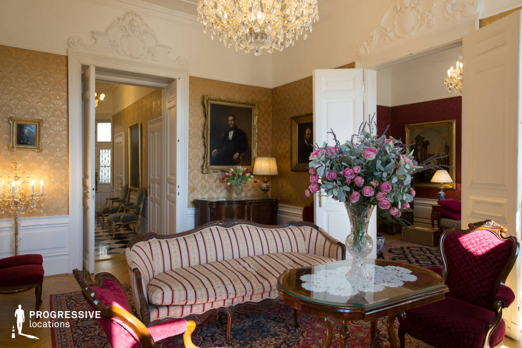 This opulent salon radiates classic European elegance, adorned with gilded wallpaper, crystal chandeliers, and finely carved white moldings. The high ceilings and tall double doors create a sense of grandeur, seamlessly connecting multiple rooms with equally rich décor. A centerpiece striped antique sofa with curved wooden detailing sits atop a Persian-style rug, surrounded by deep red velvet chairs and an ornately carved wooden table. The space is accentuated by gold-framed oil portraits, a dark wood sideboard, and an elaborate candelabra. The adjoining rooms, visible through open doors, continue the luxurious aesthetic with rich textiles and classical furnishings. This setting is ideal for period dramas, high-end editorial photography, or luxury lifestyle productions, offering a richly detailed and historically resonant ambiance.