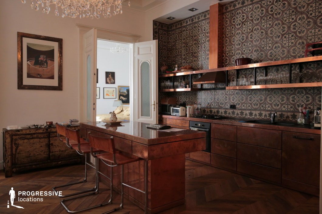 This sophisticated kitchen combines rich copper finishes with intricate patterned tiles, creating a warm and refined ambiance. The custom-built copper cabinetry and island reflect a luxurious, artisanal aesthetic, contrasted by open metal shelving that adds an industrial touch. The deep-hued wooden chevron flooring enhances the historic character of the space. A grand crystal chandelier brings a sense of elegance, complementing the high ceilings and classic white-paneled doors that lead into an adjoining room decorated with framed artwork and vintage furnishings. This setting is ideal for high-end interior photography, period film productions, or luxury lifestyle editorials, offering a visually striking blend of heritage and modern craftsmanship.