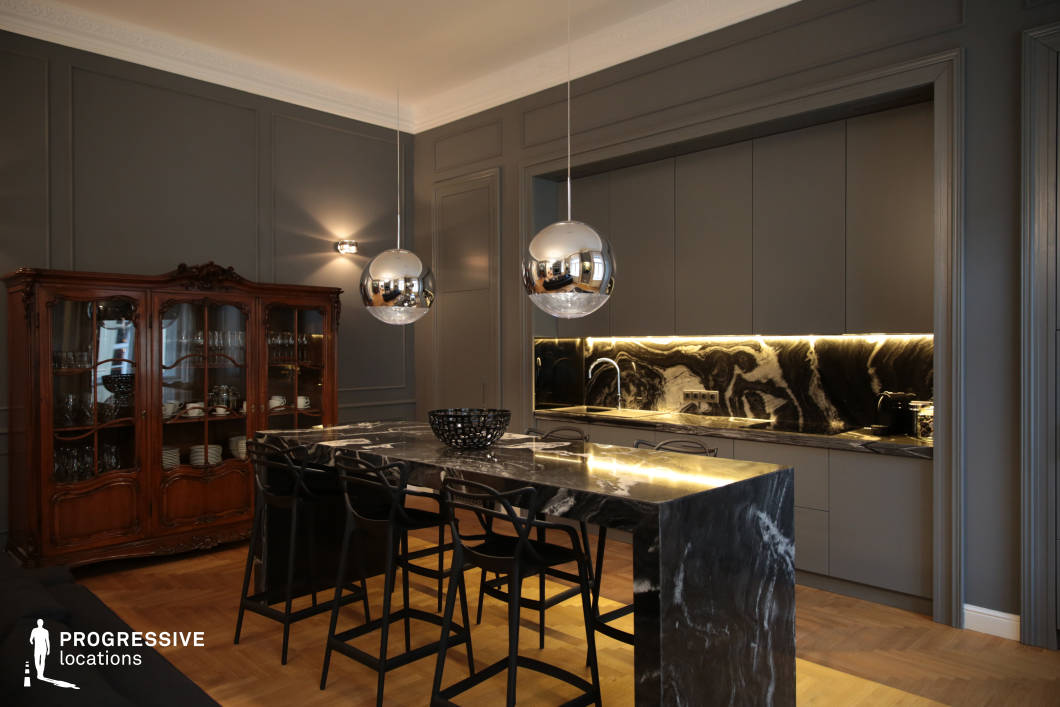 n, featuring deep gray walls, sleek cabinetry, and a dramatic black marble island with bold veining. The matching marble backsplash is illuminated by warm under-cabinet lighting, enhancing the rich textures and moody ambiance. Suspended chrome pendant lights add a contemporary touch, reflecting the room’s refined aesthetic. The space is anchored by a traditional wooden display cabinet with ornate carvings, introducing a contrast between heritage craftsmanship and minimalist modernity. The combination of dark tones, high-end materials, and soft lighting creates an intimate yet luxurious atmosphere, making this space ideal for high-end interior photography, upscale lifestyle productions, or contemporary architectural showcases.