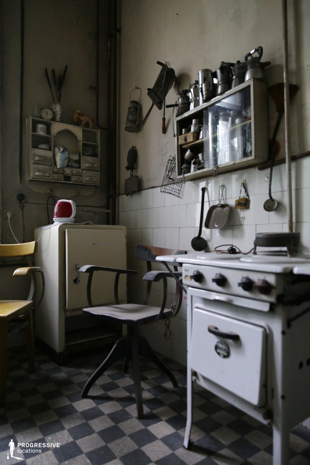 This compact vintage kitchen exudes an aged, utilitarian charm with its worn checkered linoleum flooring, tiled walls, and retro furnishings. The space is dominated by an old-fashioned white enamel stove, a compact refrigerator, and a mix of wooden and metal chairs, contributing to its eclectic and timeworn aesthetic. Open wall-mounted shelves and hanging kitchenware reinforce the industrial-era functionality. The muted lighting and visible wear on surfaces enhance the nostalgic atmosphere, making this an ideal setting for historical film productions, documentary photography, or retro-inspired editorial shoots.