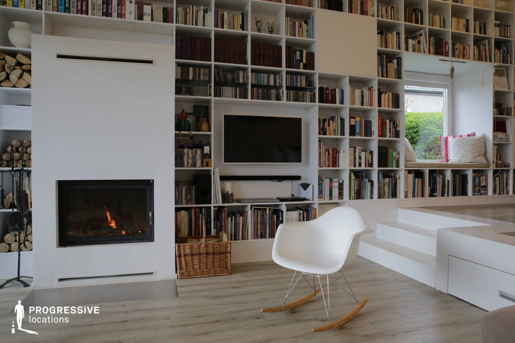 This modern living space seamlessly blends comfort and functionality, featuring a floor-to-ceiling bookshelf integrated with a sleek fireplace and media center. The predominantly white color scheme enhances the clean, minimalist aesthetic, while the warm wood flooring adds a touch of coziness. A built-in window seat provides a quiet reading nook, framed by a large bookshelf filled with a mix of literature and decorative elements. The fireplace serves as a focal point, with neatly stacked firewood reinforcing the inviting ambiance. A white rocking chair introduces a mid-century modern touch, complementing the room’s structured yet relaxed atmosphere. This space is ideal for interior photography, editorial shoots, or lifestyle productions, offering a refined yet welcoming setting.