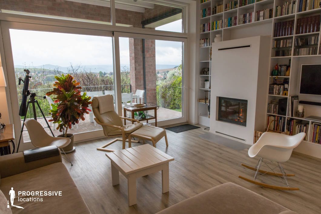This open-concept living room offers a seamless blend of indoor and outdoor spaces, featuring floor-to-ceiling glass doors that lead to a spacious terrace with scenic views of rolling hills. The interior is designed with a modern, minimalist aesthetic, incorporating warm wooden flooring, a built-in white fireplace, and an expansive bookshelf that extends to the ceiling. A combination of sleek and classic seating, including a sculptural rocking chair and a reclining lounge chair, enhances the relaxed yet sophisticated ambiance. The outdoor terrace, framed by exposed brick columns, provides an inviting extension of the living space, making this setting ideal for lifestyle photography, interior design showcases, or editorial productions that emphasize contemporary elegance and natural connectivity.