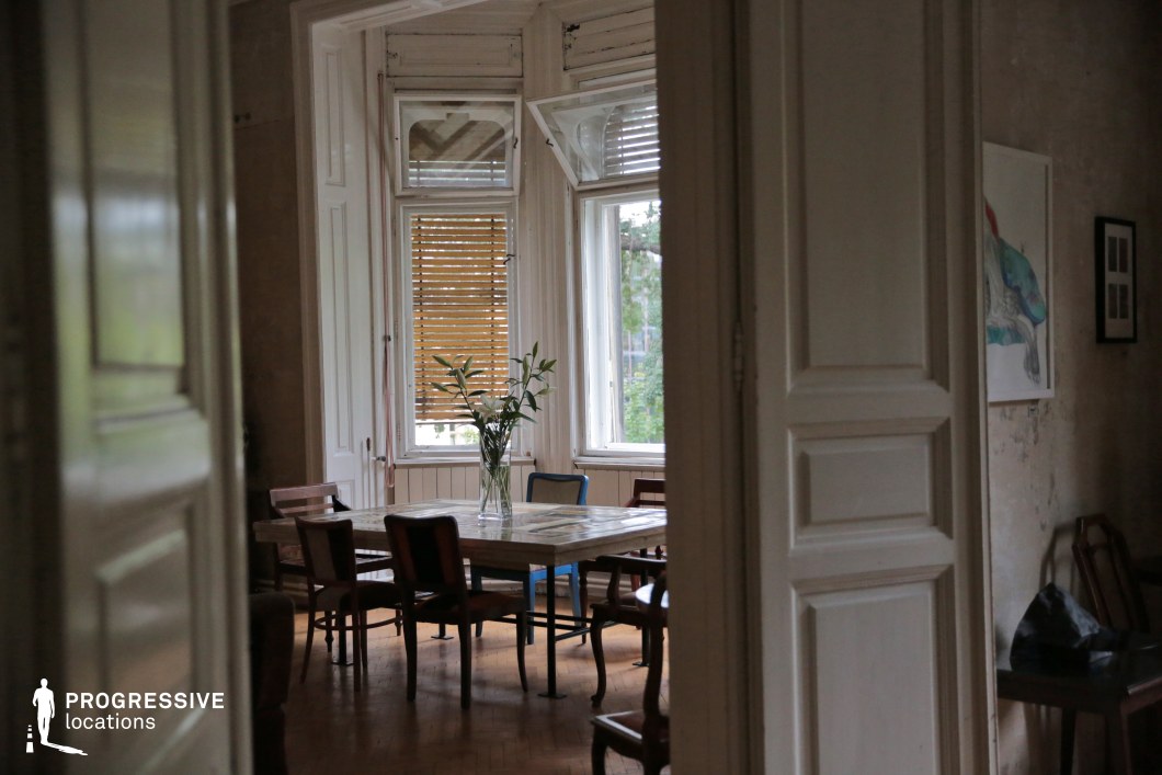 This atmospheric dining room captures a sense of quiet elegance, framed by tall, paneled double doors that open into a bright yet aged interior. Large windows with wooden shutters allow soft, diffused natural light to enter, highlighting the room’s distressed walls and classic wooden flooring. A sturdy, timeworn dining table surrounded by an eclectic mix of chairs reinforces the space’s vintage charm. The high ceilings and subtle architectural details add to the grand yet intimate ambiance. This location is ideal for artistic film settings, period photography, or editorial shoots that emphasize nostalgia and understated sophistication.