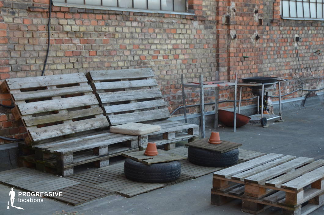 This urban rooftop terrace embraces a raw, industrial aesthetic, featuring exposed brick walls, large factory-style windows, and makeshift seating constructed from repurposed wooden pallets. The worn concrete flooring and metal shelving contribute to the rugged, unfinished atmosphere. The space is accentuated by a minimalist arrangement of upcycled materials, including wooden planks and stacked tires, reinforcing the improvised, urban feel. The open layout and industrial backdrop make this location ideal for alternative fashion shoots, gritty urban storytelling, or creative productions seeking a raw, DIY-inspired environment.