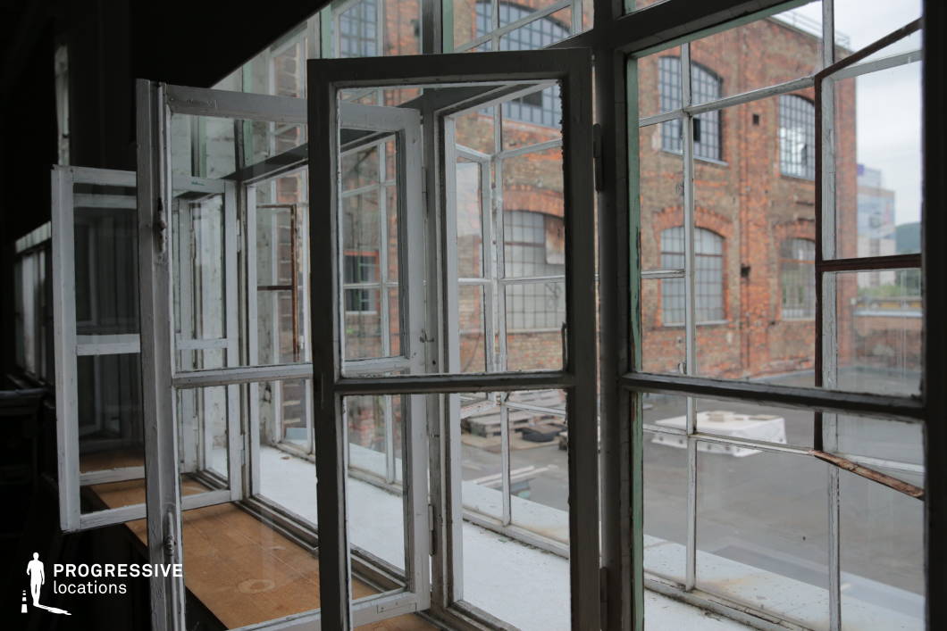 This industrial loft features large, multi-pane metal-framed windows that open outwards, offering a striking view of a historic brick factory building. The aged white-painted window frames show signs of wear, adding to the raw, vintage charm of the space. Natural light filters through the expansive glass panes, enhancing the depth and texture of the exposed brick exterior. The open windows create a dynamic interplay between the interior and the surrounding urban environment, making this an ideal setting for industrial-style photography, artistic film productions, or conceptual editorial shoots focused on architectural heritage and atmospheric depth.