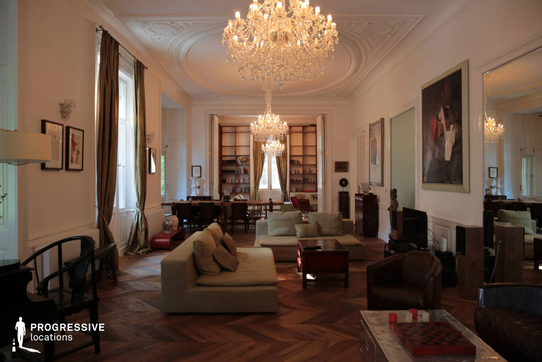 This opulent salon exudes timeless luxury with its high ceilings, intricate moldings, and crystal chandeliers that illuminate the space with a warm glow. The symmetrical layout is centered around an elegant seating arrangement featuring plush sofas and armchairs in neutral tones, contrasting with the rich wooden herringbone flooring. Tall windows framed by flowing drapery allow natural light to highlight the architectural details. The open-plan design seamlessly connects to a refined library space, complete with floor-to-ceiling bookshelves and a stately writing desk. Large mirrors and contemporary artwork blend classical and modern influences, making this setting ideal for high-end interior photography, period dramas, or editorial productions that require a sophisticated yet inviting ambiance.