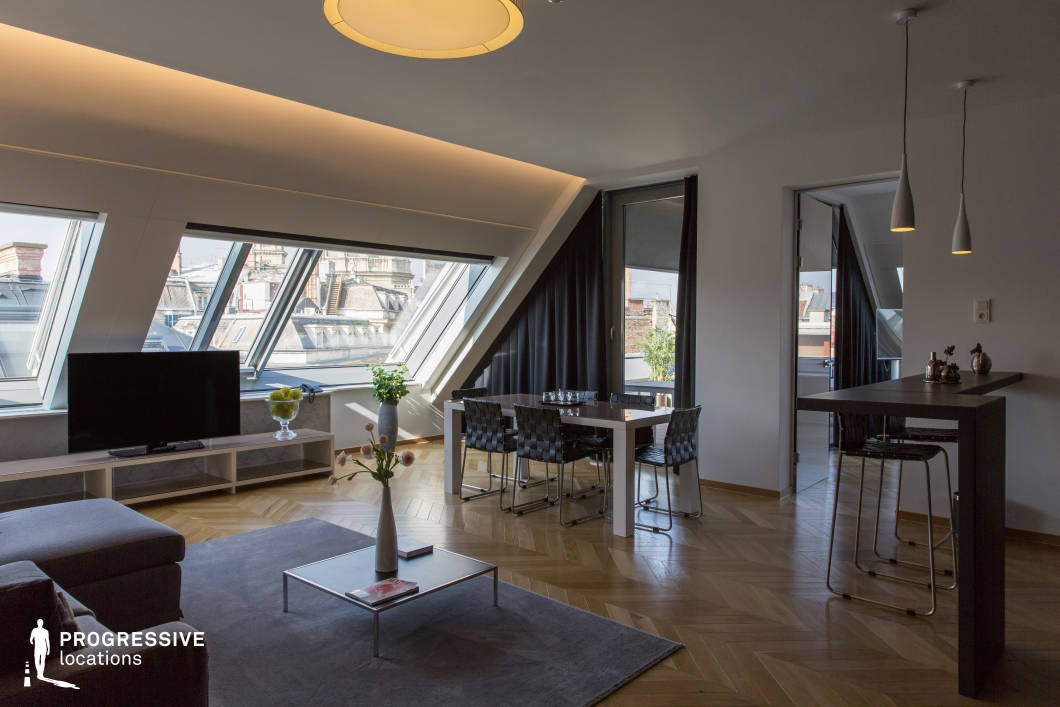 This modern penthouse living space features floor-to-ceiling angled windows that offer striking views of the surrounding rooftops, flooding the interior with natural light. The open-plan layout includes a spacious lounge area, a sleek dining space, and a contemporary high-top bar, all unified by warm herringbone wood flooring. The neutral color palette is complemented by dark furnishings and minimalist decor, creating a refined yet inviting atmosphere. Integrated recessed lighting and pendant fixtures add a sophisticated touch, while access to a private terrace enhances the connection to the urban landscape. This setting is ideal for luxury lifestyle productions, architectural showcases, or contemporary editorial photography.