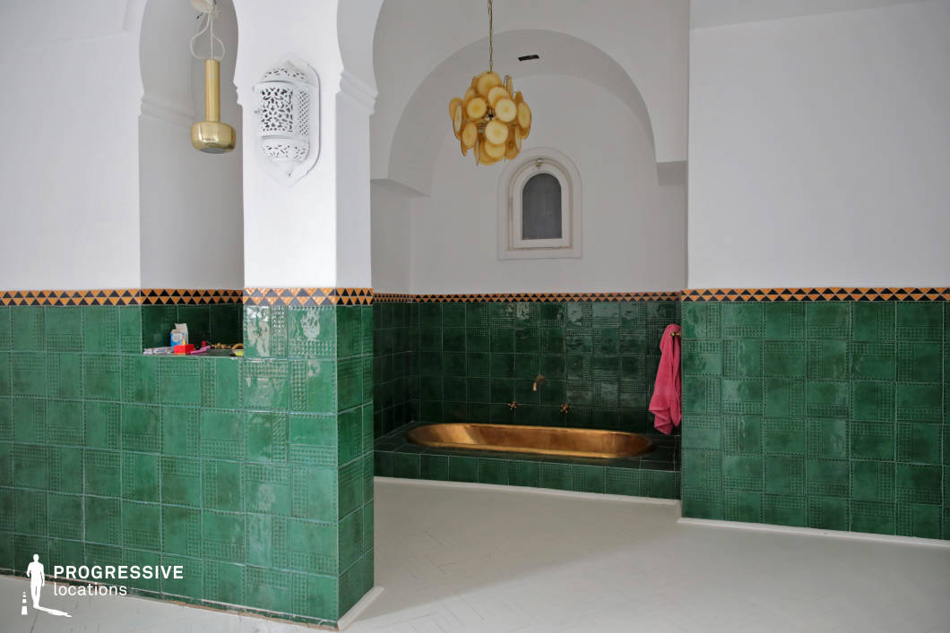 This opulent bathhouse showcases a blend of traditional Moroccan architecture and refined craftsmanship. The space is adorned with deep green glazed tiles featuring intricate geometric patterns, contrasted by crisp white walls and elegant archways. A sunken golden bathtub serves as the focal point, adding a luxurious element to the space. Ornamental wall sconces and a sculptural pendant light enhance the ambiance, reflecting the region's rich decorative heritage. The combination of smooth, polished surfaces and intricate detailing creates an immersive, tranquil atmosphere, making this setting ideal for editorial photography, luxury interior showcases, or film productions evoking timeless elegance and cultural authenticity.