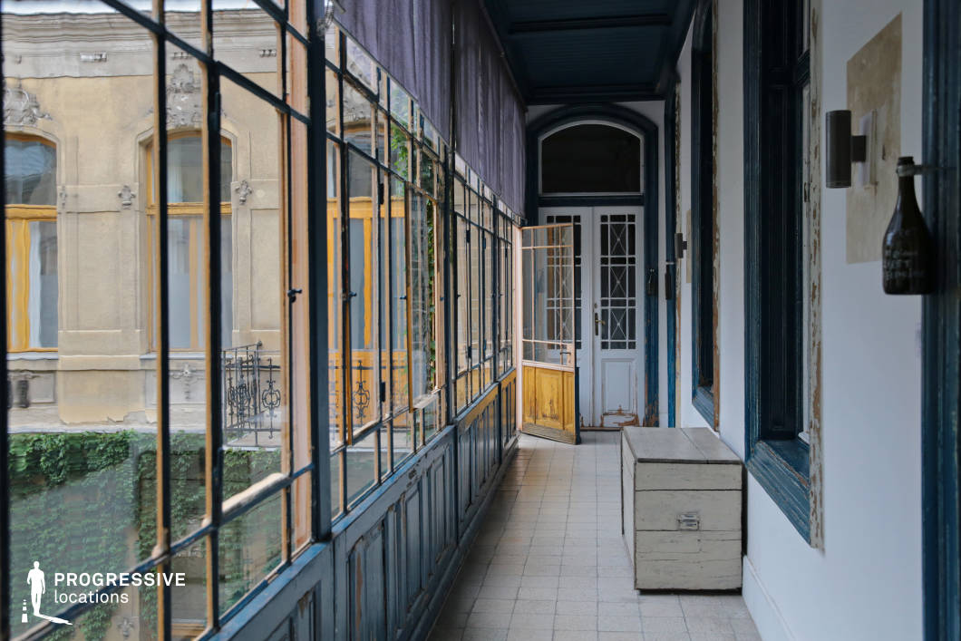This elegant covered loggia exudes old-world character, featuring large metal-framed windows that overlook a classic European courtyard. The space is framed by deep blue wooden paneling with distressed paint, adding to its aged charm. The tiled floor leads to an ornate doorway with intricate glass panels, reinforcing the historical ambiance. Across the courtyard, the facade of an adjacent building showcases decorative stucco details and warm yellow window frames, enhancing the sense of architectural heritage. This atmospheric location is ideal for period film settings, editorial photography, or projects that require a nostalgic yet sophisticated backdrop.