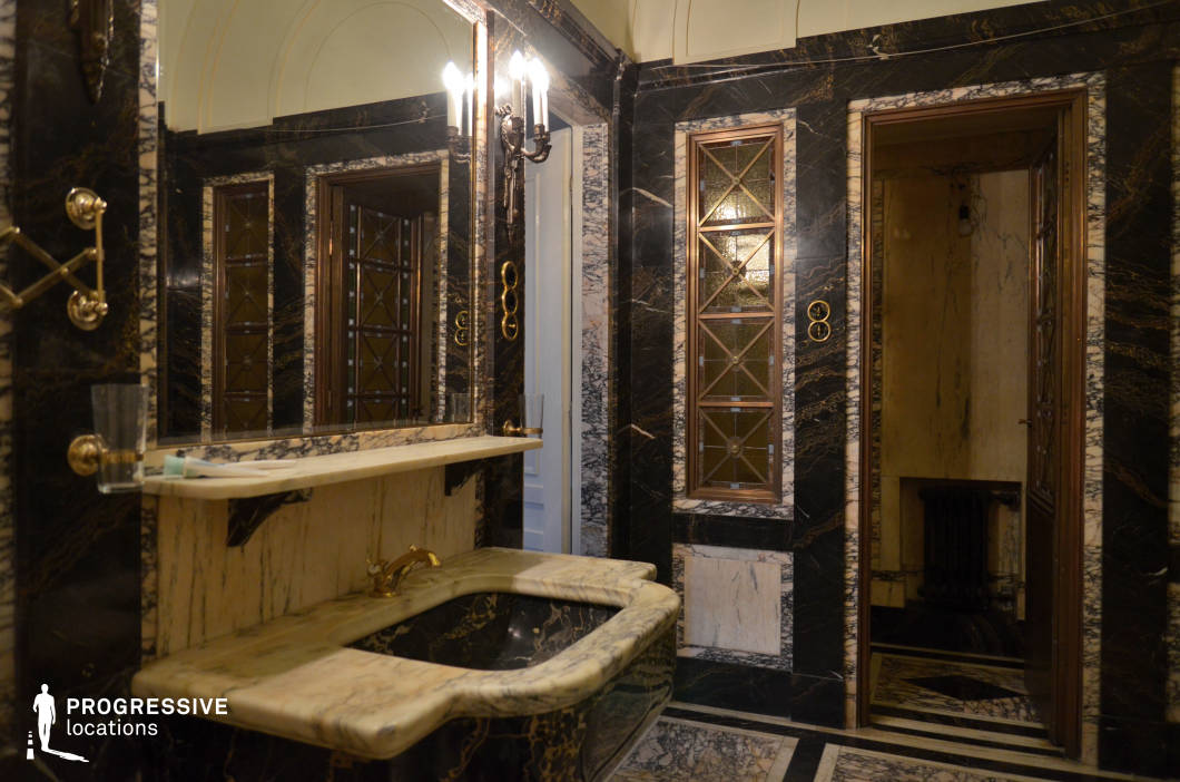 This opulent bathroom showcases a striking combination of black and cream marble, exuding timeless sophistication. The space features intricate gold-accented fixtures, geometric stained-glass panels, and a sculptural marble sink seamlessly integrated into the walls. A large mirror reflects the ambient glow from elegant wall sconces, enhancing the depth and grandeur of the room. Architectural details, including arched ceilings and finely crafted wooden doors, contribute to the historic charm. This setting is ideal for luxury interior photography, period film productions, or high-end editorial shoots that require a rich, dramatic atmosphere.