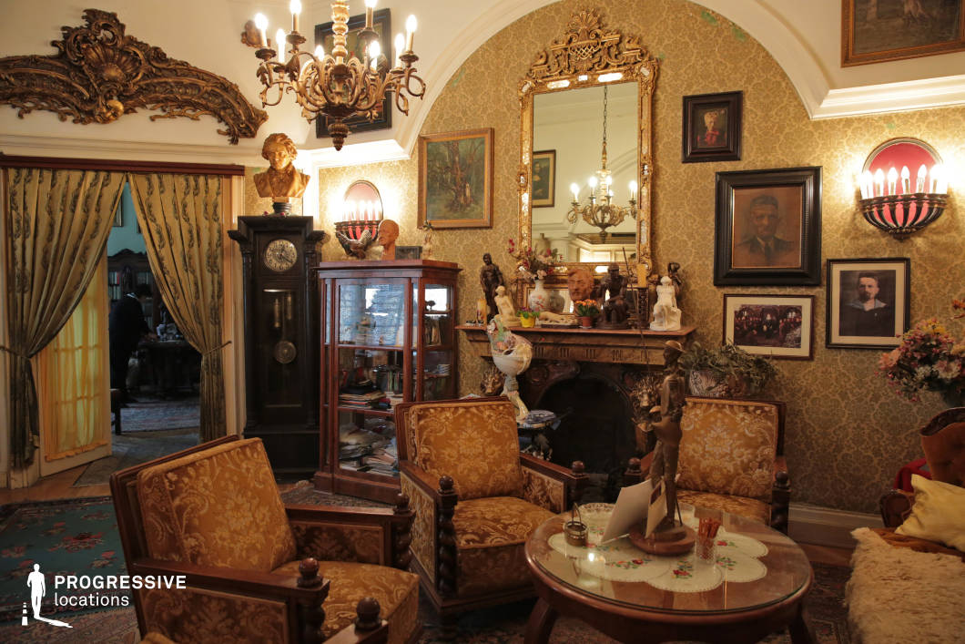 This richly decorated salon exudes old-world elegance, featuring intricate gold-accented wallpaper, carved wooden furnishings, and a grand fireplace framed by an opulent gilded mirror. The high, arched ceiling is adorned with decorative moldings and an elaborate chandelier, adding to the room’s grandeur. Heavy draped curtains separate adjoining rooms, enhancing the stately ambiance. Classic upholstered armchairs with intricate wooden detailing and an antique grandfather clock contribute to the historic character. Ideal for period dramas, editorial photography, or productions requiring a luxurious, vintage European setting.
