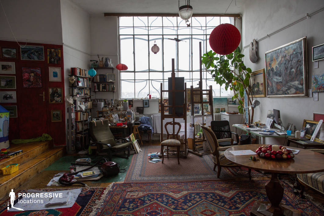 This artist’s studio features an expansive industrial-style window with intricate metal framing, allowing abundant natural light to illuminate the space. The high ceilings enhance the openness, while white walls provide a neutral backdrop for an eclectic collection of paintings and books. The floor is covered with richly patterned Persian rugs that add warmth and texture. Various seating options, including vintage wooden and upholstered chairs, contribute to the inviting, lived-in ambiance. The studio is characterized by its mix of vintage furniture, layered textiles, and a creative, bohemian atmosphere, making it an ideal setting for film scenes requiring a working studio, an intellectual retreat, or a space with timeless European charm.