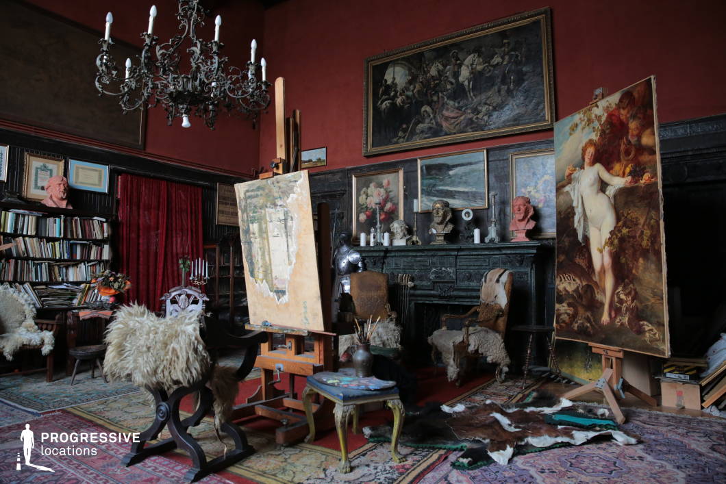 This artist’s studio features a richly decorated, baroque-inspired interior with dark wooden paneling, deep red walls, and an elaborate wrought-iron chandelier that enhances the historical ambiance. The space is filled with large-scale classical paintings, ornate furniture, and antique decorative elements that contribute to its dramatic and opulent character. An intricately carved fireplace serves as a focal point, flanked by framed artwork and sculptures, adding to the grandeur of the setting. The floor is covered with layered Persian rugs and animal hides, reinforcing the vintage and aristocratic atmosphere. The studio’s unique combination of historical charm and artistic functionality makes it an ideal location for period films, classic portrait photography, or settings requiring a sophisticated, old-world aesthetic.