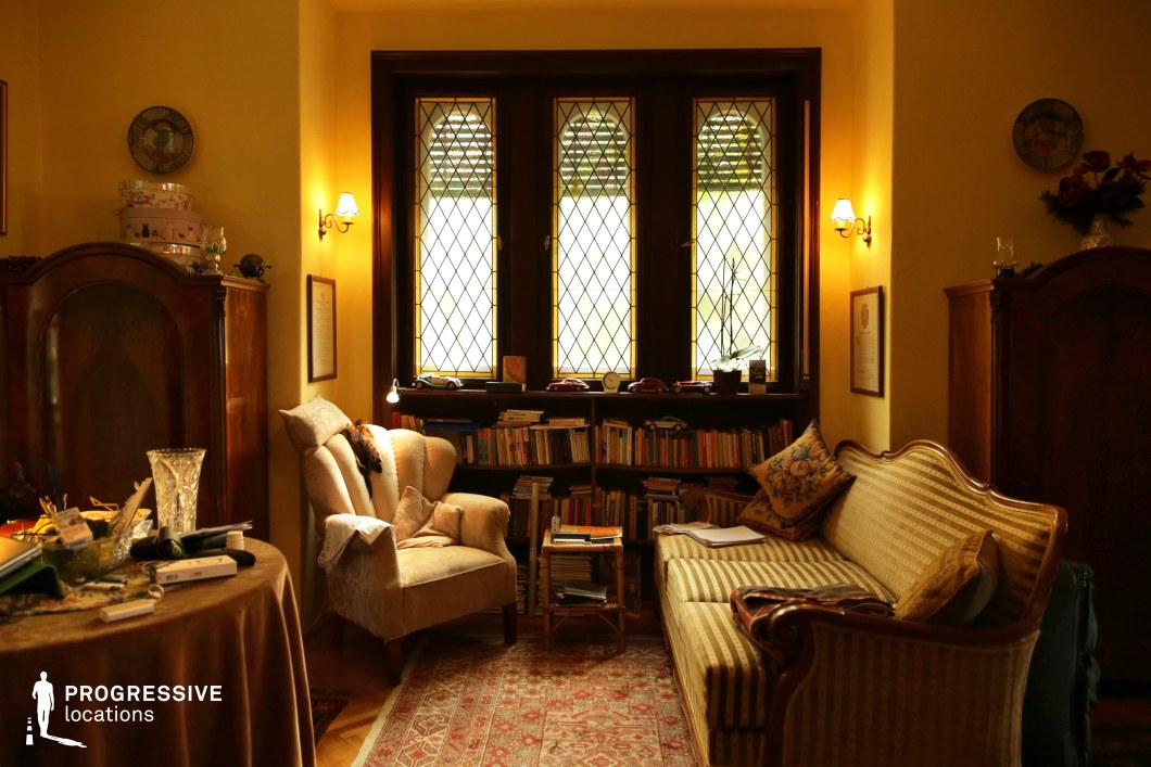 This intimate reading room exudes a vintage charm with its warm yellow walls, classic wooden furnishings, and soft ambient lighting. The space is anchored by a large window with diamond-patterned glass panes, framed in rich dark wood, which enhances the old-world aesthetic. A plush armchair and an elegant striped sofa create a comfortable and inviting atmosphere, complemented by an extensive built-in bookshelf filled with well-worn volumes. Ornate wooden cabinets and traditional decorative details reinforce the room’s historical character, while the patterned rug adds texture and warmth. This refined yet cozy setting is ideal for film scenes requiring a nostalgic, intellectual, or literary ambiance, as well as editorial and period-piece photography.