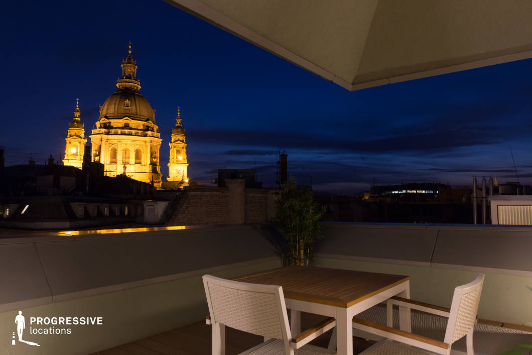 This modern rooftop terrace features a sleek, minimalist design with clean architectural lines and neutral tones that enhance its sophisticated aesthetic. The open layout provides an unobstructed, dramatic view of a grand historic basilica, creating a striking visual contrast between contemporary and classical architecture. The refined furnishings and subtle greenery add a touch of elegance, seamlessly integrating with the urban surroundings. The elevated positioning and carefully curated design make this an ideal location for high-end lifestyle photography, cinematic scenes, or exclusive rooftop events.
