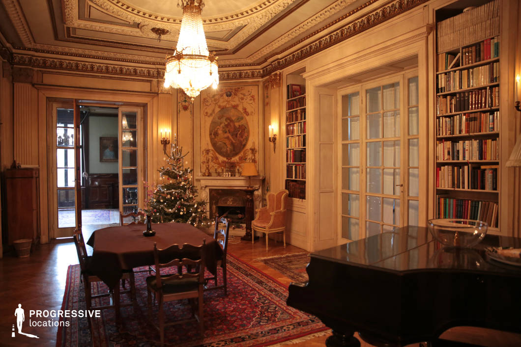 This refined classical salon showcases intricate architectural detailing, with decorative moldings, gilded accents, and a coffered ceiling that enhances its grandeur. A crystal chandelier serves as the focal point, casting a warm glow over the richly furnished space. The built-in bookshelves, ornate fireplace, and large double doors framed by elegant wood paneling contribute to the room’s timeless sophistication. A grand piano and antique furnishings add to the aristocratic ambiance, while the richly patterned rugs and delicate wall paintings reinforce the historical charm. This opulent yet inviting setting is ideal for period film productions, luxury lifestyle photography, or scenes requiring an atmosphere of classical refinement and intellectual prestige.