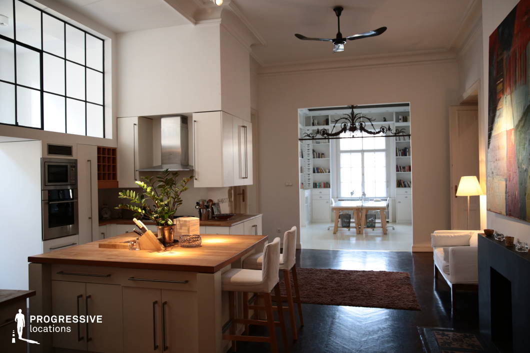 This spacious open-concept interior seamlessly blends modern functionality with classic architectural elements, featuring high ceilings, large industrial-style windows, and a neutral color palette. The kitchen is defined by sleek cabinetry, wooden countertops, and a central island that enhances both aesthetics and practicality. The dark polished flooring contrasts elegantly with the bright white walls, creating a sophisticated yet inviting atmosphere. An adjacent study area, framed by built-in bookshelves and a large window, adds a refined touch, making the space feel both expansive and intimate. The combination of clean lines, soft lighting, and carefully curated furnishings makes this an ideal setting for lifestyle photography, upscale home interiors, or film scenes requiring a stylish yet livable ambiance.