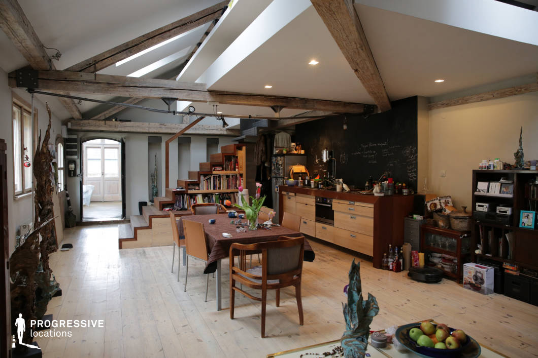 This spacious loft seamlessly blends industrial and rustic design elements, featuring exposed wooden beams, slanted ceilings, and a mix of raw materials that create a warm yet modern aesthetic. The open floor plan enhances the sense of space, with a functional kitchen area, a central dining space, and a built-in staircase leading to an elevated section with integrated bookshelves. Light wood flooring contrasts with darker furniture tones, while large windows and skylights allow for abundant natural illumination. The combination of rough-hewn textures, sleek cabinetry, and an open-concept layout makes this an ideal setting for creative workspaces, artistic residences, or film and photography productions requiring a contemporary yet organic atmosphere.