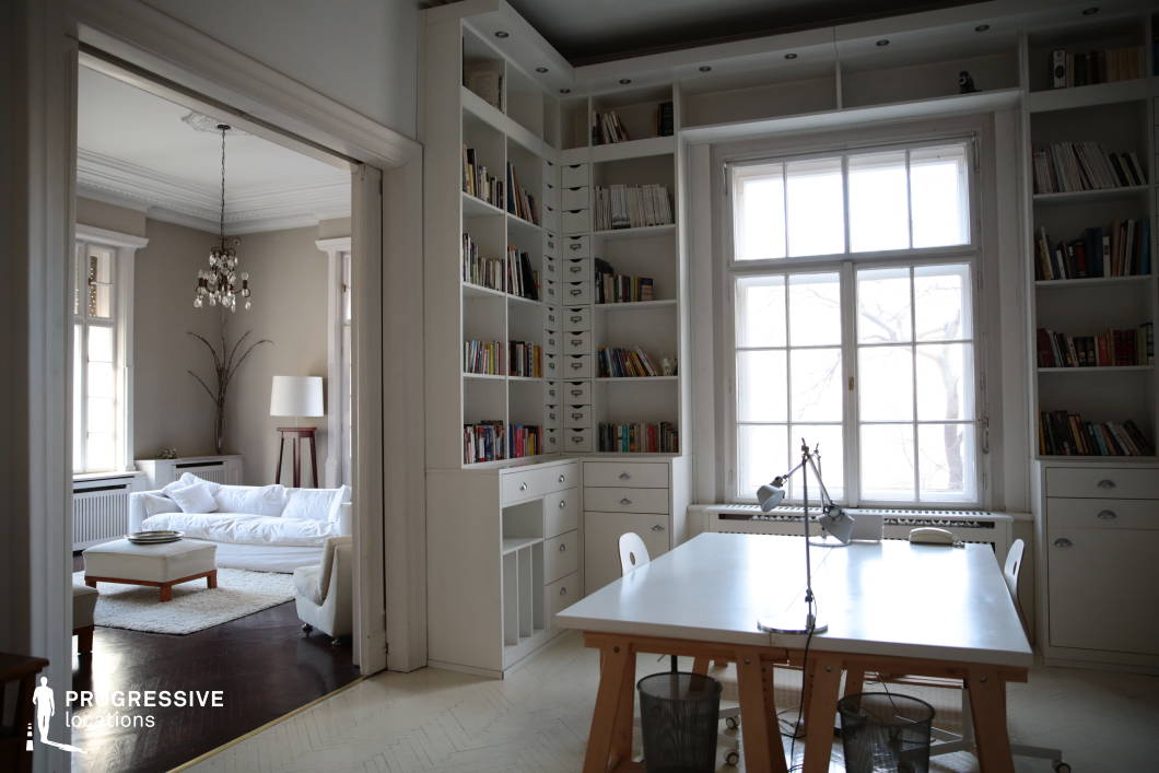 This sophisticated study seamlessly blends classic architectural details with a modern minimalist aesthetic, featuring high ceilings, intricate moldings, and large windows that create a bright and airy atmosphere. Floor-to-ceiling built-in bookshelves provide a structured yet elegant focal point, while the crisp white color palette enhances the sense of openness and refinement. The adjoining living area, visible through a wide doorway, continues the cohesive design with soft furnishings, dark wood flooring, and a statement chandelier, balancing warmth with understated luxury. The combination of functional workspace and stylish relaxation area makes this an ideal setting for film productions, editorial photography, or high-end interior design showcases.