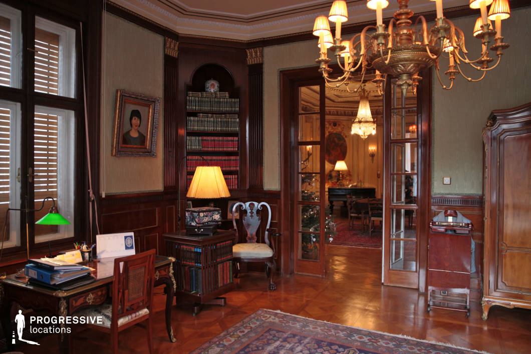 This refined study exudes a timeless sophistication, featuring rich wood-paneled walls, intricate crown moldings, and a grand chandelier that enhances the room’s classical charm. Built-in bookshelves filled with leather-bound volumes frame the space, while elegant French doors lead to an adjoining salon, seamlessly blending the study with the grandeur of the larger residence. The parquet flooring, antique furniture, and warm lighting contribute to the room’s stately ambiance, making it an ideal setting for period film productions, high-end editorial photography, or interiors requiring a distinguished and aristocratic aesthetic.