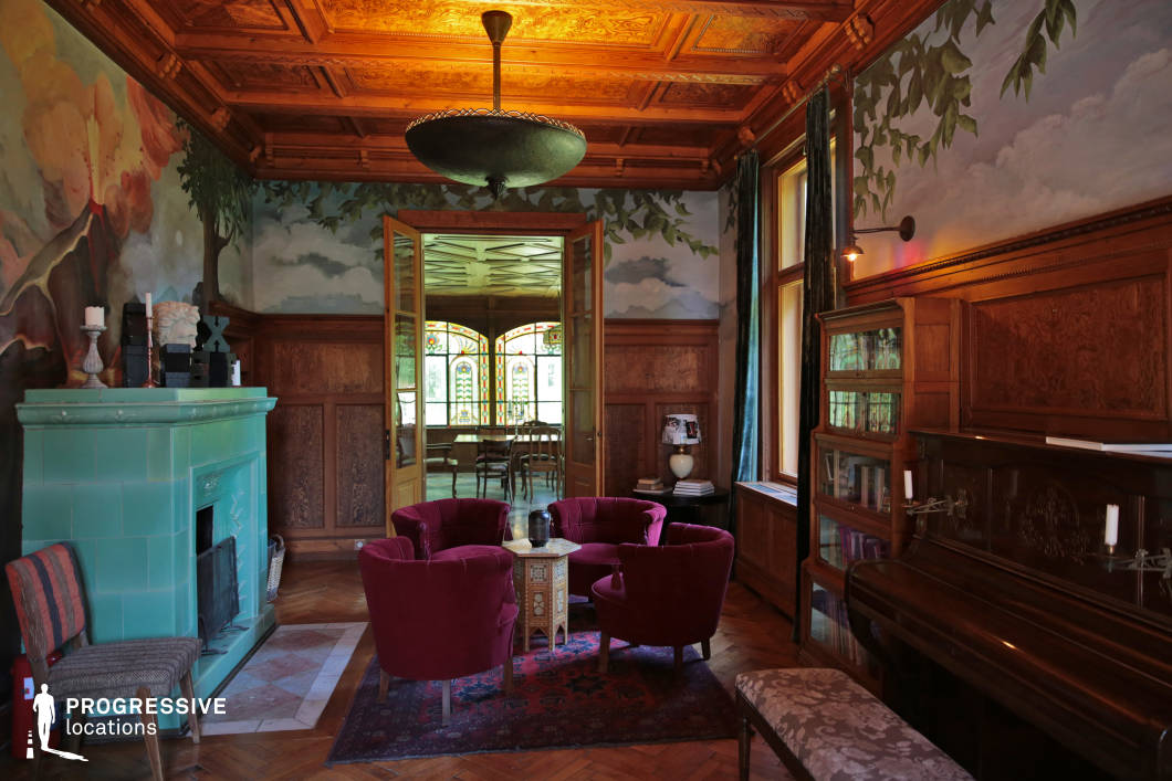 This richly detailed salon blends classic craftsmanship with artistic elegance, featuring a coffered wooden ceiling, intricate wall paneling, and hand-painted nature-inspired murals that create an immersive atmosphere. A striking green ceramic stove serves as a focal point, complementing the warm wood tones and plush furnishings. The space is framed by large windows with heavy velvet drapery, allowing soft natural light to highlight the fine details of the interior. Through elegant double doors, an adjoining room with stained-glass windows enhances the historical charm. The harmonious mix of textures, colors, and refined materials makes this an ideal setting for period film productions, luxury editorial shoots, or sophisticated interior design showcases.