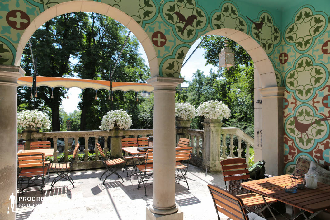 This elegant villa terrace features grand stone arches, intricate hand-painted wall motifs, and a beautifully aged balustrade overlooking lush greenery. The architectural details blend classical and decorative elements, creating a refined yet inviting atmosphere. The patterned ceiling and vibrant wall designs contrast with the natural stone flooring, while the arched openings frame the surrounding landscape, enhancing the sense of openness. The harmonious mix of historical elegance and relaxed charm makes this an ideal setting for period film productions, luxury lifestyle photography, or scenes requiring a romantic European ambiance.