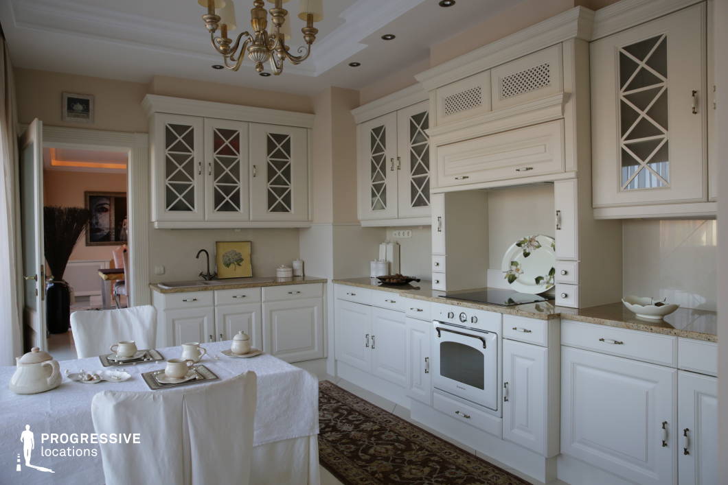 This refined kitchen embodies timeless elegance with its all-white cabinetry, intricate molding details, and a harmonious blend of traditional and contemporary design elements. The glass-paneled upper cabinets and ornate brass chandelier add a touch of sophistication, while the soft neutral tones create a bright and inviting ambiance. The layout is both functional and aesthetically pleasing, featuring built-in cabinetry, a vintage-style oven, and an elegant dining area with coordinated furnishings. The seamless connection to an adjoining living space enhances the sense of openness, making this an ideal setting for high-end interior photography, lifestyle productions, or film scenes requiring a sophisticated and serene kitchen backdrop.