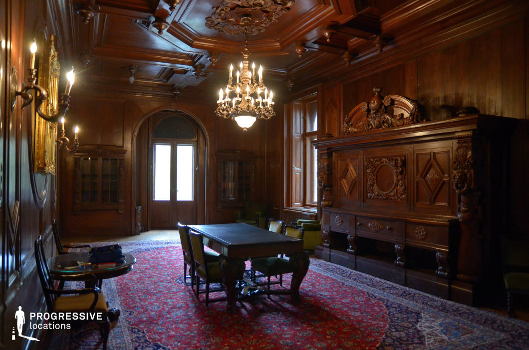 This grand, wood-paneled study exudes a timeless elegance with its intricately carved walls, coffered ceiling, and rich, dark wood tones. A large central table surrounded by upholstered chairs anchors the space, sitting atop an ornate, deep-colored rug that enhances the room’s stately atmosphere. The carved wooden cabinetry and an elaborately detailed fireplace contribute to the sense of historical grandeur, while the arched double doors and tall windows allow for a balanced interplay of natural light. A chandelier and wall-mounted sconces provide warm, ambient illumination, emphasizing the craftsmanship and depth of the space. This refined setting, with its classical architectural elements and rich textures, is ideal for sophisticated period settings or high-profile executive interiors.