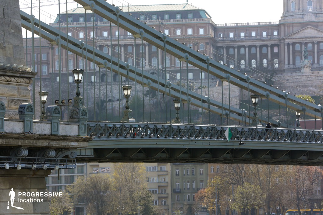 This location image captures a close-up view of a historic suspension bridge with ornate ironwork and decorative lampposts, set against the monumental backdrop of a grand neoclassical palace. The bridge structure features a repetitive rhythm of vertical cables and diagonal steel beams, creating a layered, geometric composition. The iron railings are detailed with lattice patterns, and the lampposts are mounted on sculpted bases that reflect 19th-century design aesthetics. The dominant color palette includes muted grays and greens of the bridge structure, harmonizing with the earth-toned stone and brick of the background architecture. The textures of wrought iron, weathered stone, and aged masonry add a rich historical character to the scene. The large-scale palace behind the bridge establishes a powerful architectural presence with its symmetrical façade, arched windows, and classical proportions. The image composition emphasizes depth through the overlapping structural lines of the bridge and the receding architectural layers. This location offers a visually compelling, urban historical setting that is ideal for productions requiring a majestic cityscape with a strong sense of European heritage.