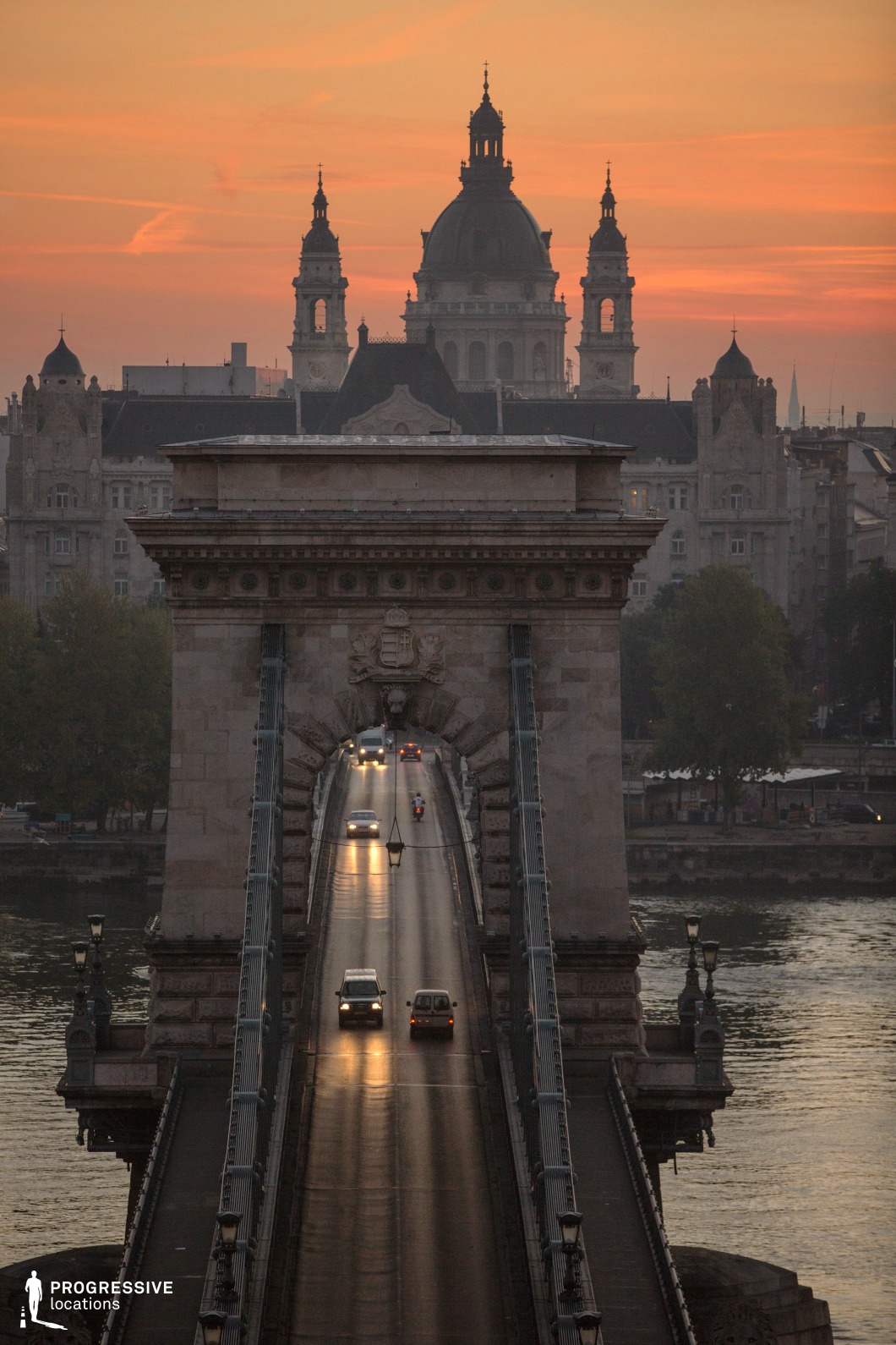 This image captures a historic suspension bridge with a monumental stone gateway, leading the eye towards a grand domed building in the background. The bridge's massive stone pillars and ornate iron suspension cables highlight its 19th-century engineering, while the decorative lampposts along the walkway enhance its classical elegance. The repetitive linear elements of the suspension cables and roadway converge towards the architectural focal point in the distance, creating a strong sense of depth and perspective. The background features an imposing neoclassical building with intricate detailing and symmetrical towers, reinforcing the city's historical character. The bridge serves as a dramatic frame, guiding visual focus toward the architectural landmark beyond. This location is ideal for cinematic urban compositions, period-piece settings, or showcasing European architectural grandeur within a structured cityscape.