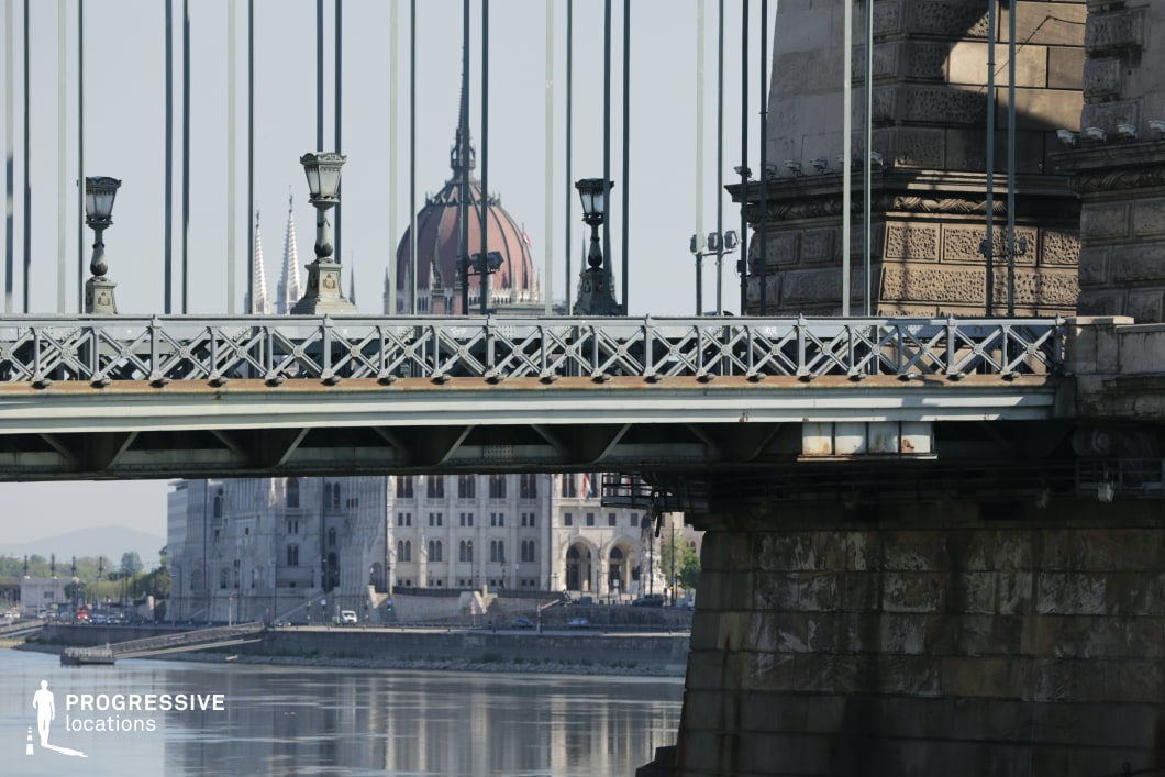 This image presents a detailed view of a historic suspension bridge, emphasizing its ornate iron railings, decorative lampposts, and massive stone foundation. The intricate lattice design and vertical suspension cables create a structured, rhythmic composition, showcasing 19th-century engineering with a refined industrial aesthetic. The massive stone pillar anchoring the bridge features intricate masonry work, adding a sense of durability and craftsmanship. In the background, a grand neogothic-style building with a prominent dome and spires is framed through the bridge’s structural elements. This architectural layering enhances the depth of the composition and establishes a strong visual contrast between the bridge’s industrial ironwork and the ornate historical facades beyond. The scene is well-suited for cinematic cityscapes, architectural documentation, or period-set urban storytelling, highlighting the relationship between infrastructure and monumental city landmarks.