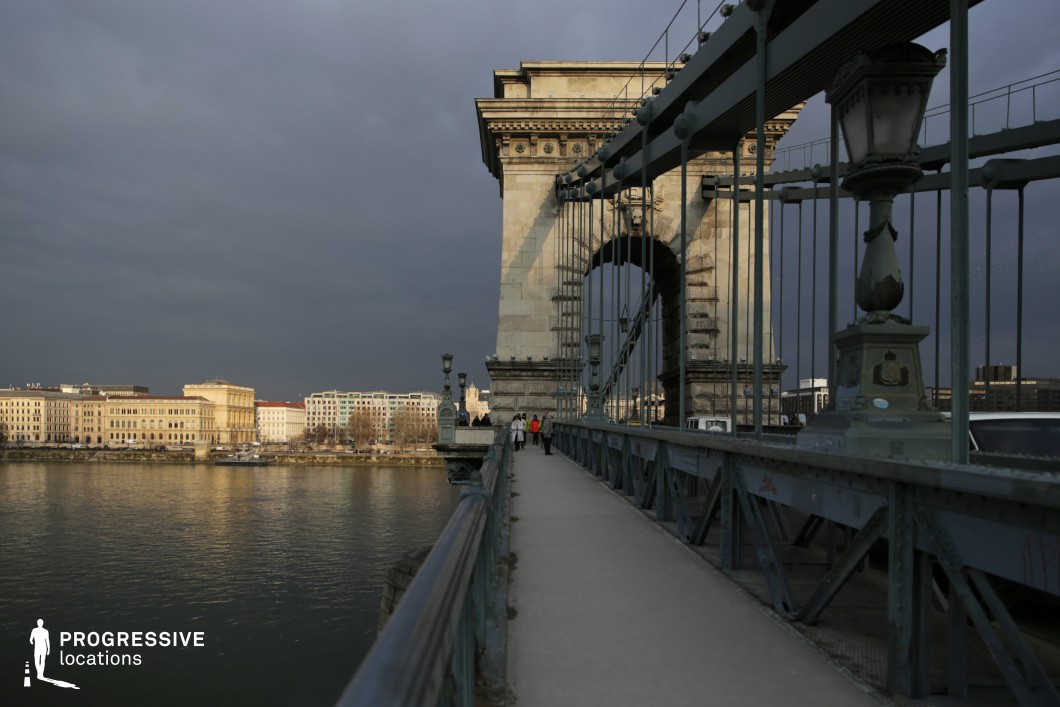This image captures a historic suspension bridge with a grand stone archway, leading towards a riverside cityscape. The bridge features intricate metal railings, vertical suspension cables, and ornate lampposts, which emphasize its 19th-century engineering and classical architectural influences. The pedestrian walkway runs alongside the road, providing a strong leading perspective that guides the eye toward the monumental gateway. In the background, a row of elegant historic buildings with symmetrical facades and large windows lines the riverbank, adding depth and architectural contrast. The massive stone structure anchoring the bridge reinforces its solidity, while the intricate detailing of the lampposts and decorative elements adds refinement. This location is ideal for architectural cinematography, period-set urban photography, and cityscape compositions, showcasing the relationship between historic infrastructure and a well-preserved European riverside setting.