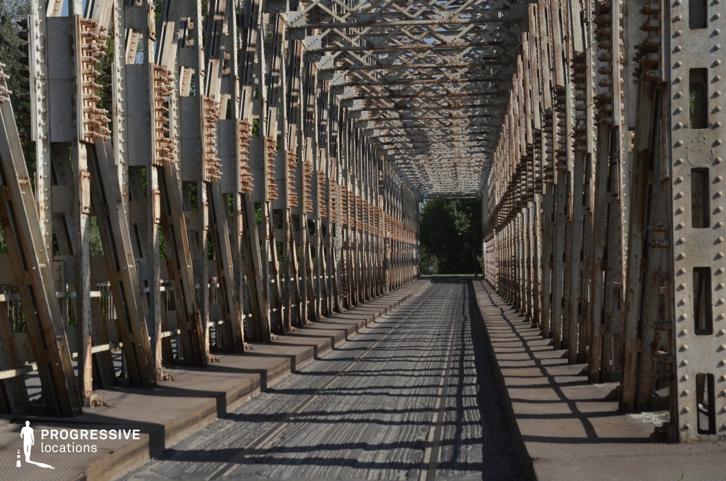 This image captures a narrow metal truss bridge, featuring a repetitive lattice framework that creates a striking tunnel-like effect. The bridge’s riveted steel beams and diagonal braces form a complex geometric pattern, emphasizing its industrial engineering style. The metal surfaces show signs of weathering, adding a sense of age and history to the structure. The long, enclosed perspective leads the viewer’s eye toward a tree-lined opening at the far end, contrasting the rigid metal framework with the natural environment beyond. The strong lines and shadows cast by the beams enhance the depth and rhythm of the composition. This location is ideal for industrial-themed cinematography, architectural photography, or conceptual urban visuals, where structured repetition and historical infrastructure play a key role in the scene’s aesthetics.