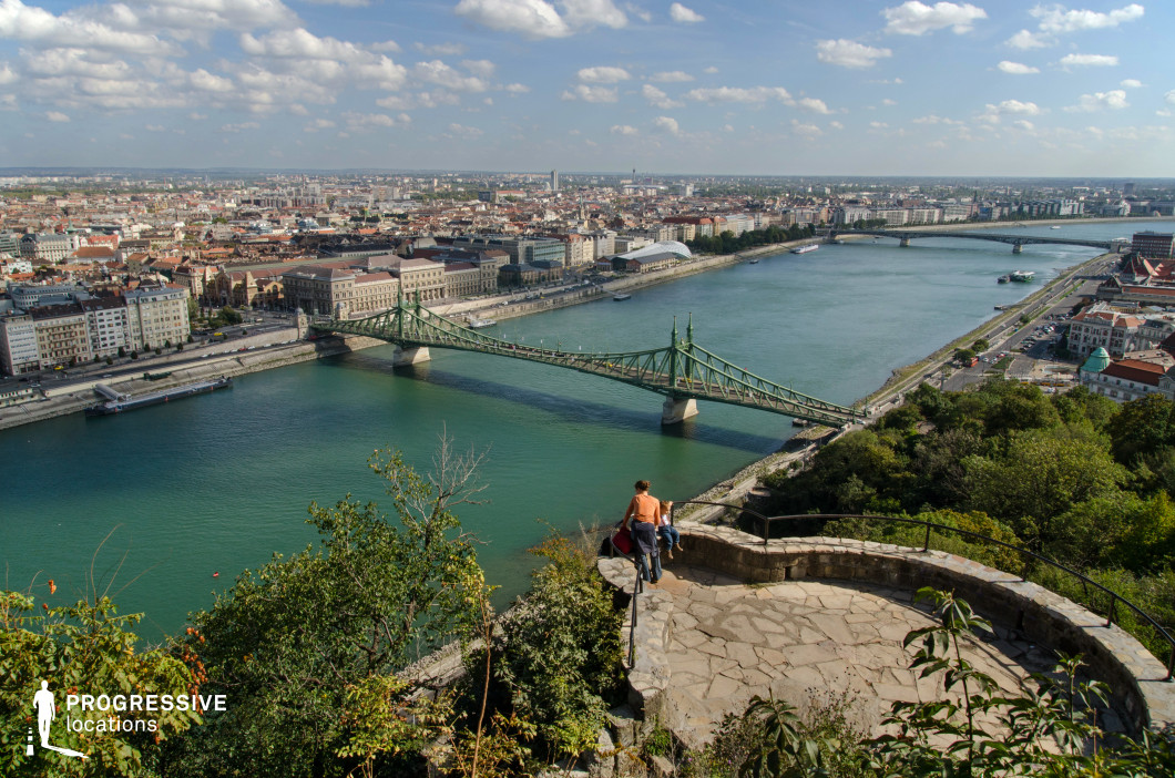This image presents a stunning panoramic view of a historic European city, captured from an elevated stone terrace with a curved railing. The scene overlooks a green iron truss bridge spanning a wide river, connecting two urban districts. The bridge’s ornate metal framework and sturdy stone piers reflect classic industrial-era engineering, harmonizing with the surrounding neoclassical and modern architecture along the riverbanks. In the foreground, lush greenery and rugged stone pathways add a natural contrast to the structured cityscape below. The river, cutting through the city, provides depth, with additional bridges and historic buildings visible in the distance. This location is ideal for cinematic cityscape shots, urban exploration photography, and travel-related visual storytelling, capturing the fusion of historic infrastructure and natural vantage points.