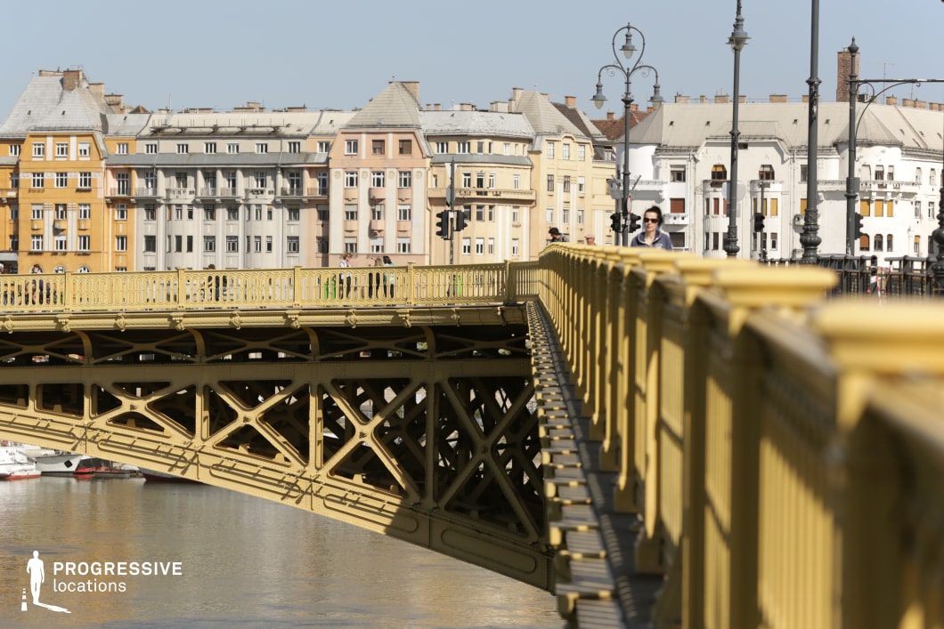 This image features a historic yellow iron bridge, showcasing intricate lattice metalwork, decorative railings, and elegant lampposts. The bridge’s ornate architectural details and rhythmic repetition of railings create a visually dynamic composition. Its riveted metal structure and graceful arches emphasize 19th-century engineering aesthetics, blending both strength and elegance. In the background, a row of historic European buildings with colorful facades and classic architectural details forms a vibrant urban setting. Their varied heights, window styles, and warm color palette contrast with the structured precision of the bridge. The scene is ideal for cinematic urban compositions, architectural photography, or visual storytelling focused on historic infrastructure and city life, capturing the relationship between classic European architecture and industrial-era engineering.