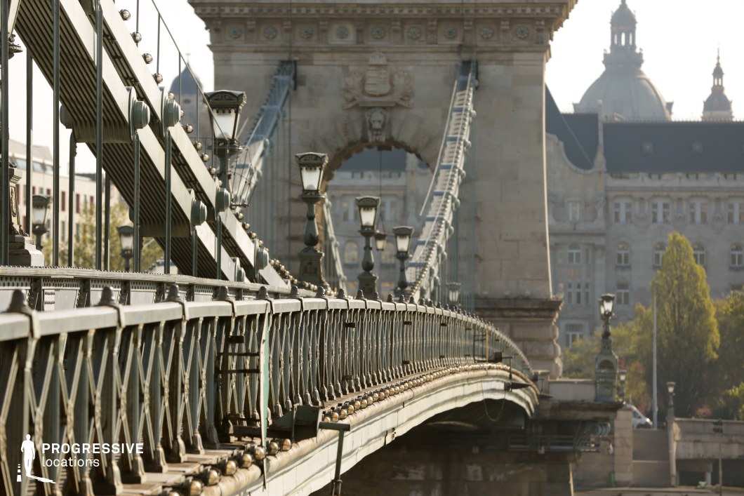 This image highlights a historic suspension bridge, featuring intricate iron railings, elegant lampposts, and riveted structural details. The sweeping curve of the bridge guides the eye towards the massive stone archway, adorned with classical decorative elements. The repeating vertical suspension cables and lampposts create a strong rhythmic perspective, enhancing the bridge’s grand, industrial-era aesthetic. In the background, a stately historic building with graceful arched windows, and another historic domed building, complement the bridge’s refined architectural style, reinforcing the scene’s European urban character. The interplay of ornate iron craftsmanship and monumental stonework makes this location ideal for architectural cinematography, historical cityscape photography, and period-set urban visuals, capturing the elegance and engineering mastery of 19th-century infrastructure.