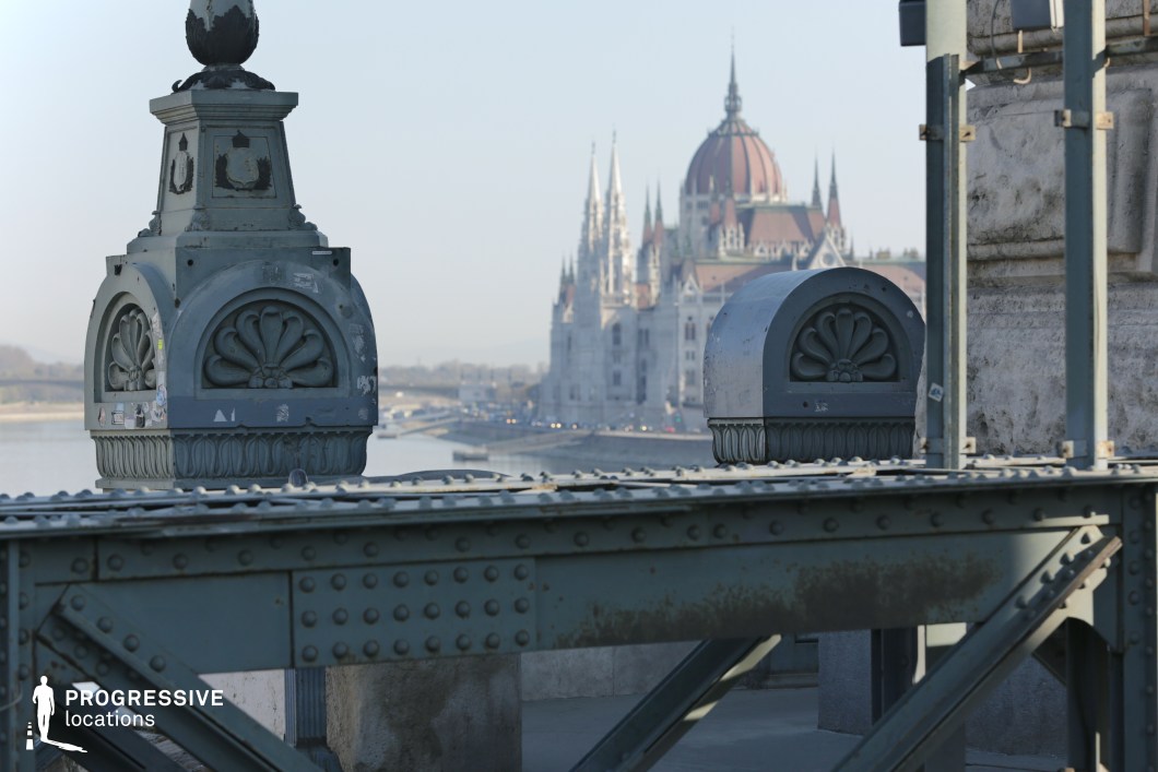 This image focuses on the intricate decorative ironwork of a historic bridge, showcasing finely crafted floral motifs, riveted metal elements, and ornamental structures. The detailed cast-iron elements exhibit a blend of industrial engineering and classical decorative influences, characteristic of 19th-century European bridge design. The weathered metal surfaces add a sense of history and durability to the structure. In the background, the grand neo-Gothic Parliament building with pointed spires and a central dome rises above the river, reinforcing the scene’s architectural grandeur. The contrast between the bridge’s industrial iron framework and the Parliament’s elaborate stone façade creates a striking juxtaposition. This location is ideal for architectural cinematography, historical cityscape photography, and urban visual storytelling, emphasizing the interplay of industrial infrastructure and monumental architecture.