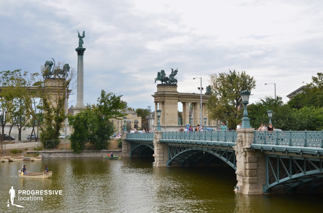 This image showcases a decorative historic iron bridge with elegant ornamental railings, intricate lampposts, and arched metal supports. The bridge’s cast-iron detailing and stone foundations reflect classical European engineering, blending aesthetic refinement with structural stability. The soft blue-green hue of the ironwork enhances its timeless charm, contrasting with the natural water and greenery surrounding it. In the background, monumental stone structures adorned with statues, columns, and bronze equestrian sculptures dominate the scene, reinforcing the grand, historical ambiance of the location. The presence of trees and open water creates a harmonious balance between urban and natural elements. This setting is ideal for historical cityscape cinematography, architectural photography, and visual storytelling, capturing the elegance of classical European infrastructure alongside monumental heritage landmarks.
