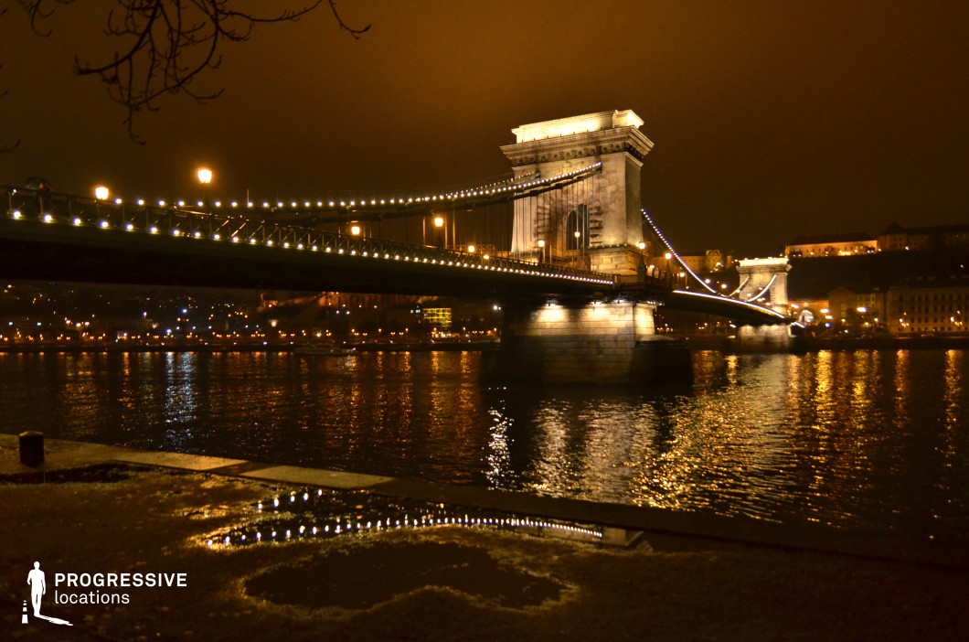 This image showcases a grand historic suspension bridge, elegantly illuminated against the night sky. The bridge's string lights trace the suspension cables, creating a visually striking arc that emphasizes its architectural symmetry and structural elegance. The massive stone pillars, bathed in warm light, provide a sense of solidity and permanence, anchoring the composition. The reflections of the golden city lights shimmer on the water’s surface, adding depth and movement to the scene. In the background, a historic hilltop complex with softly lit facades enhances the bridge’s majestic presence. The combination of warm artificial lighting and deep shadows makes this setting ideal for cinematic night cityscapes, atmospheric urban photography, and visual storytelling that highlights historic European landmarks.