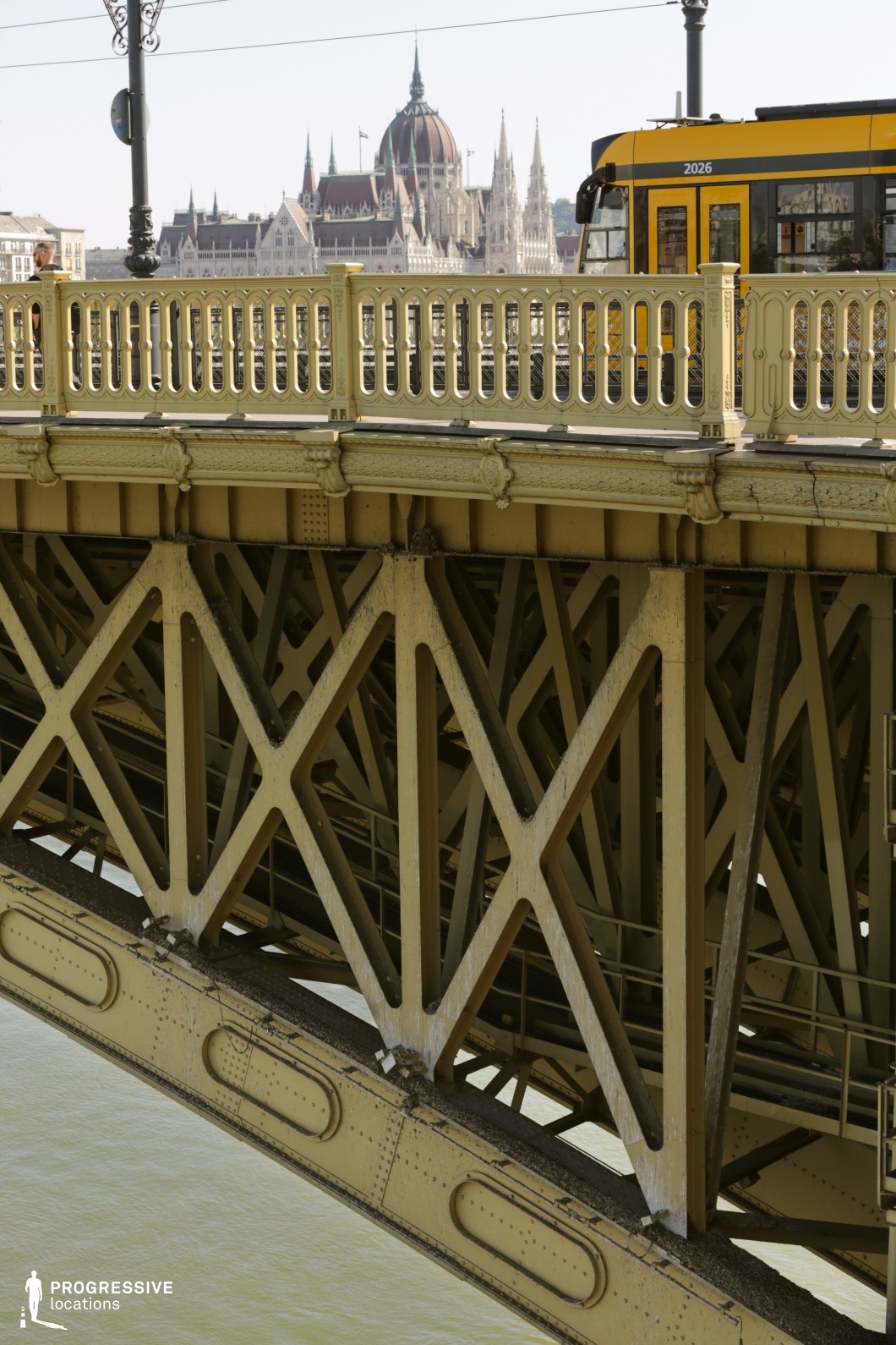 This image captures a historic yellow iron bridge, highlighting its intricate lattice framework, decorative railings, and riveted steel supports. The bold geometric patterns of the trusses emphasize the bridge’s industrial-era engineering, while the finely detailed cast-iron balustrades add a touch of elegance. The contrast between the solid lower structure and the refined upper railings enhances the visual depth of the composition. In the background, the grand neo-Gothic Parliament building with soaring spires and a distinctive dome stands as a striking focal point, reinforcing the city’s historical architectural richness. The presence of a yellow tram moving across the bridge adds a dynamic urban element, emphasizing the bridge’s active role in public transportation. This location is ideal for cinematic cityscape shots, architectural photography, and urban storytelling, blending historic infrastructure with contemporary movement and European architectural grandeur.