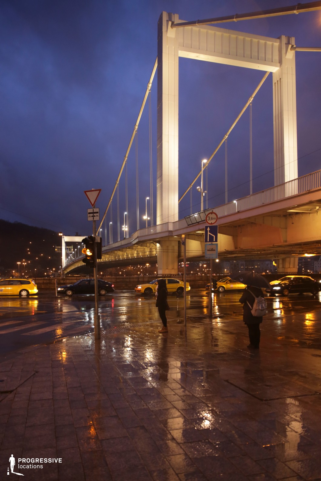 This image showcases a modern white suspension bridge with geometric pylons and taut suspension cables, forming a sleek and contemporary structure. The bridge’s artificial illumination contrasts sharply with the deep blue evening sky, emphasizing its minimalist and functional design. The evenly spaced streetlights and clean architectural lines enhance its structured aesthetic. In the foreground, a busy city intersection with traffic signals and pedestrian crossings reinforces the bridge’s urban connectivity. The combination of modern infrastructure and dynamic city elements makes this location ideal for cinematic cityscapes, urban architectural photography, and modern infrastructure-focused storytelling.