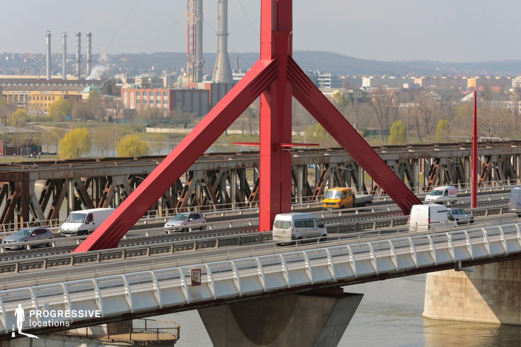This image showcases a modern cable-stayed bridge, distinguished by its bold red steel pylons and geometric structural design. The angled support beams and sleek white railing system contribute to its contemporary aesthetic, while the multi-lane roadway emphasizes its role as a major transportation route. The bridge's sturdy concrete foundation and metal framework provide a strong sense of stability. In the background, an industrial landscape with smokestacks, factories, and mixed urban architecture creates a contrasting setting between modern infrastructure and functional city outskirts. An older, weathered steel railway bridge running parallel to the main road adds another layer of structural depth. This location is ideal for cinematic industrial cityscapes, transportation infrastructure photography, and urban development visuals, highlighting the juxtaposition of modern engineering and industrial heritage.
