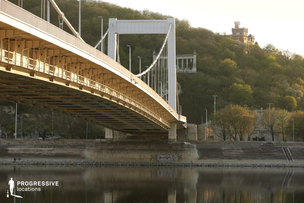 This image captures a modern white suspension bridge, elegantly spanning a river with its clean geometric lines and cable-supported structure. The bridge’s steel framework and suspension cables create a sense of lightness, contrasting with the massive stone piers anchoring it to the riverbanks. The warm sunlight highlights the intricate metalwork beneath the roadway, emphasizing its structural details. In the background, a green hillside dotted with classical monuments and a historic brick tower provides a striking contrast to the modern infrastructure. The natural setting blends seamlessly with urban development, creating a harmonious balance between contemporary engineering and historic architecture. This location is ideal for cinematic cityscapes, architectural photography, and infrastructure-focused visuals, capturing the intersection of modern transportation and cultural heritage.