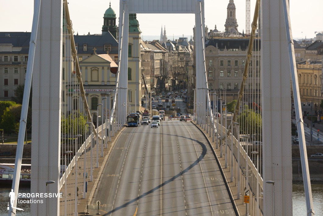 This image captures a modern white suspension bridge, its sleek pylons and taut cables framing the view of an elegant urban boulevard. The bridge’s clean architectural lines and symmetrical structure guide the eye towards the historic cityscape beyond, creating a compelling sense of depth and movement. The evenly spaced streetlights and suspension cables reinforce the bridge’s structured aesthetic, emphasizing its role as a vital transportation artery. Beyond the bridge, the dense urban setting showcases a mix of classical and baroque architecture, with ornate facades, domed towers, and decorative details contributing to the city’s rich historical character. The contrast between the bridge’s contemporary engineering and the surrounding heritage buildings makes this scene ideal for cinematic urban compositions, architectural photography, and visual storytelling about the intersection of modern infrastructure and historical city planning.