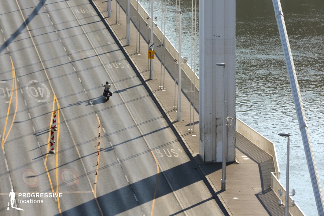 This image highlights a modern white suspension bridge, emphasizing its clean architectural design and structured symmetry. The towering steel pylons and taut suspension cables frame the bridge’s expansive roadway, creating a dynamic perspective. The road markings, barriers, and evenly spaced streetlights contribute to the bridge’s functional and urban aesthetic. The interplay of light and shadow on the road surface enhances the visual depth, while the adjacent pedestrian walkway follows the bridge’s streamlined geometry. The river below provides a natural contrast, reinforcing the balance between infrastructure and the surrounding environment. This location is ideal for cinematic cityscapes, urban infrastructure photography, and modern transportation visuals, showcasing the precision and scale of contemporary bridge engineering.