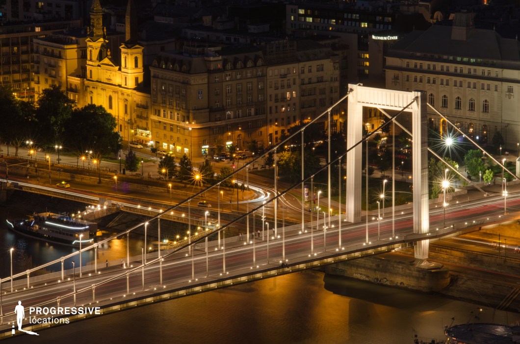 This image showcases a modern white suspension bridge, gracefully spanning across the river with elegant lighting that highlights its clean architectural lines. The illuminated streetlights and suspension cables create a rhythmic perspective, while the reflections on the water add depth and atmosphere. In the background, historic buildings with warm golden lighting contrast with the contemporary structure of the bridge, enhancing the blend of old and new urban aesthetics. The curved roadways and intersecting streets introduce a dynamic element, emphasizing the city's bustling nighttime energy. This setting is ideal for cinematic urban visuals, architectural night photography, and modern infrastructure storytelling, offering a striking balance between historical charm and contemporary engineering.