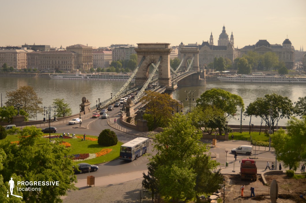 The image showcases a grand suspension bridge with imposing stone pillars and iron cables stretching across a wide river, seamlessly connecting two parts of the city. The structure is characterized by its symmetrical design and intricate ironwork, reflecting 19th-century engineering. The entrance to the bridge features ornate street lamps and decorative elements, enhancing its historic elegance. The foreground presents a curved roundabout encircled by manicured greenery, structured flowerbeds, and a paved pedestrian pathway leading towards the bridge. The surrounding urban environment is composed of neoclassical and baroque-style buildings, exhibiting detailed facades and large windows, which contribute to the old-world charm of the location. The river reflects the city's architecture, adding depth and dynamism to the composition. The elevated perspective offers a sweeping view of the bridge's structural alignment with the cityscape beyond, where domed rooftops and spires punctuate the skyline. The photograph captures the contrast between the monumental bridge and the softer, organic elements of the park and river, making it a visually compelling location with cinematic potential.